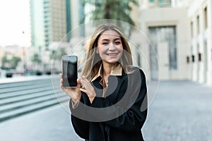 Front view of a happy business woman showing a blank smart phone display and looking to the camera in the street