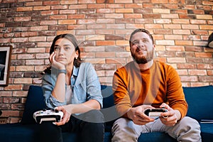 Front view of happy cheerful young couple holding controllers and playing video games on console sitting together on