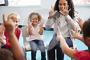 Front view of infant school children sitting on chairs in a circle in the classroom, holding up their hands and learning to count