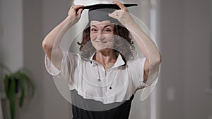 Front view intelligent proud mature woman putting on graduation cap smiling looking at camera. Medium shot portrait of