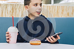 Teenager boy sits at table in cafe, drinks milkshake, eats donut, holds smartphone in his hand. Boy plays mobile games.