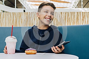 Teenager boy sits at table in cafe, drinks milkshake, eats donut, holds smartphone in his hand. Boy plays mobile games.