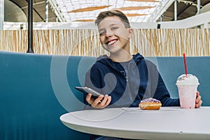 Teenager boy sits at table in cafe, drinks milkshake, eats donut, holds smartphone in his hand. Boy plays mobile games.