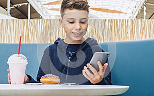 Teenager boy sits at table in cafe, drinks milkshake, eats donut, holds smartphone in his hand. Boy plays mobile games.