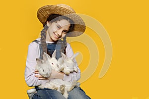 Front view of a little girl in a jeans and straw hat holding a real three white rabbits,  yellow background.