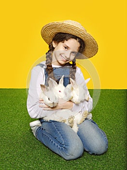 Front view of a little smiling girl in a jeans and hat holding a real three white rabbits, isolated yellow background.