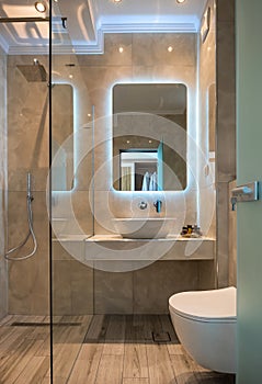 Front view of modern beige marble bathroom with vessel bowl sink, mirror back light, glass shower cabin, wall hung toilet, wooden