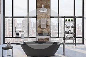 Front view on modern black bath in big sunlit loft style bathroom with dark metallic furniture, round mirror on brick wall and