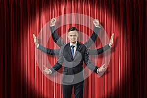 Front view of multiarmed businessman raising hands like Shiva, standing lit up by spotlight against red stage curtain.