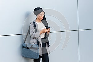 Front view of a muslim woman wearing a headscarf and holding her purse standing using her phone