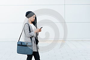 Front view of a muslim woman wearing a headscarf and holding her purse standing using her phone