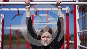 Front view portrait of blond young woman making effort doing pullups in sunbeam outdoors. Motivated Caucasian