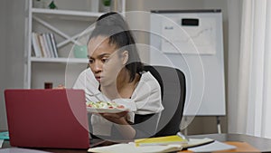 Front view portrait of busy young focused woman eating healthful veggie salad surfing Internet on laptop in home office