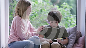 Front view portrait of Caucasian boy sitting on windowsill writing as girl scaring sibling. Frightened bullied brother