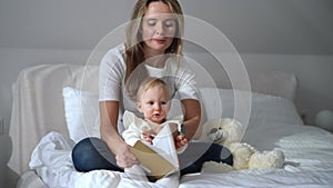 Front view portrait of Caucasian mother reading book out loud for infant daughter sitting in comfortable white bed in