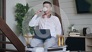 Front view portrait of cheerful handsome young Asian man drinking morning coffee in living room surfing Internet on