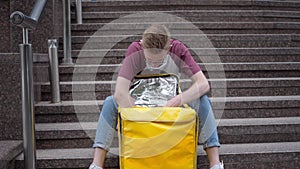 Front view portrait handsome courier unzipping yellow food delivery bag thinking sitting on urban stairs outdoors. Wide