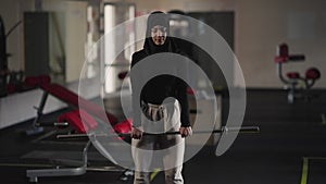 Front view portrait of motivated woman in hijab bending forward with barbell in gym. Serious confident Middle Eastern