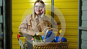 Front view portrait of young woman talking on smartphone standing with cart at storage room in yellow warehouse. Medium