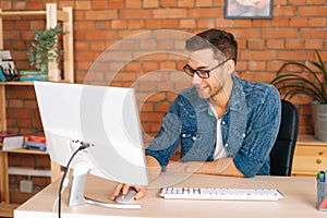Front view of positive young business man in stylish glasses working on desktop computer sitting at desk at home office