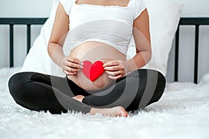 Front view of pregnant woman sit on white bed and hold red heart symbol on her belly or womb with concept love baby from mother