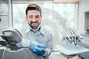 Front view of professional male dentist in white doctor coat and protective gloves sitting in dental chair and equipment, looking