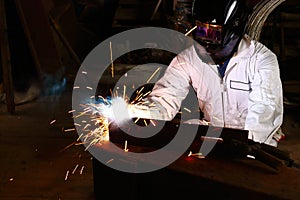 Front view of professional welder in white uniform with protective helmet welding steel with spark in workshop. Industrial worker