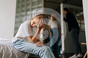 Front view of sad lonely little girl looking away, crying suffering sitting on sofa during parents quarrelling and