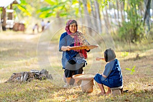 Front view of senior Asian woman with traditional clothes winnow rice using basketry and little girl stay beside and also work