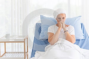 Front view of senior breast cancer patient wearing a headscarf laying in bed and praying in the hospital room, cancer concept