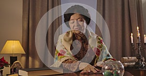 Front view of senior Caucasian woman in shawl sitting at the table with candles and shuffling cards. Positive old witch