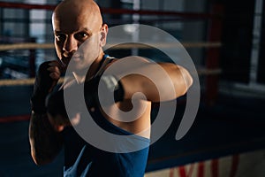 Front view of serious aggressive boxer male wearing bandages punching air to camera in sport club with dark interior, on