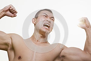Front view of shirtless, angry, roaring young man flexing his muscles with arms raised and looking away
