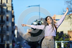 Front view of smiling beautiful young woman arms raised and standing in the street while looking camera in a sunny day