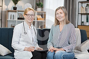 Front view of smiling mature female nurse or doctor, sitting on the sofa with her patient at home, pleasant senior lady