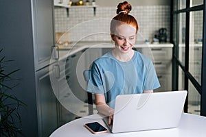 Front view of smiling redhead young woman working typing on laptop computer sitting at table.