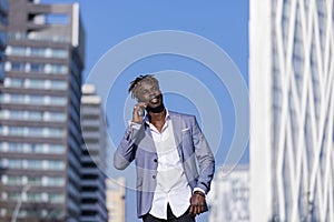 Front view of smiling young black african man wearing elegant clothes standing in the street while using a mobile phone outdoors