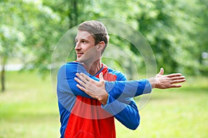 Front view of a sportsman stretching hands while standing outdoor in a park and looking by side.
