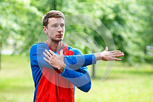 Front view of a sportsman stretching hands while standing outdoor in a park and looking by side. Close up cropped photo.