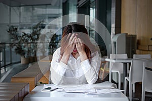 Front view of stressed frustrated young Asian business woman covering face with hands on the desk in office