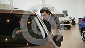 Front view of stylish car dealer in business suit talking to man client sitting in car salon. Professional salesman