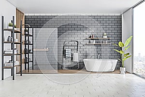 Front view on stylish sunlit bathroom with white bath, light grey concrete floor, shower on wooden wall, grey brick wall