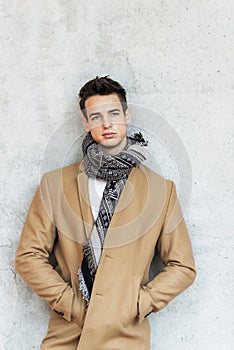 Front view of stylish young man wearing coat and scarf leaning on a wall while looking away