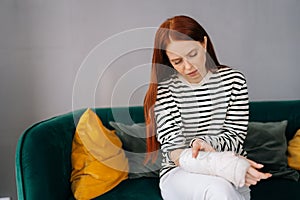 Front view of suffering from pain injured young woman with broken right hand wrapped in white gypsum bandage sitting on