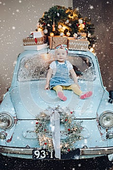 Front view of sweet and fashionable little cute girl sitting on blue retro car decorated for Christmas.