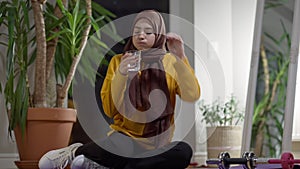Front view of tired satisfied slim fit sportswoman in hijab drinking refreshing water sitting at home after workout