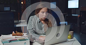 Front view of tired young woman typing on her laptop late at night. Stressed female employee using her personal computer