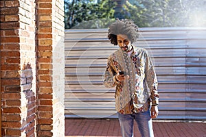 Front view of a cool young smiling afro man using a mobile phone while standing outdoors in a sunny day