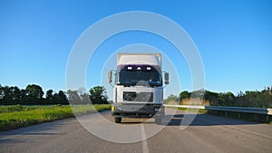 Front view of truck with cargo trailer driving on highway transporting goods at summer day. White lorry riding through