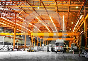 Front view of truck loading steel coil under overhead crane inside orange warehouse. Steel industry handling.